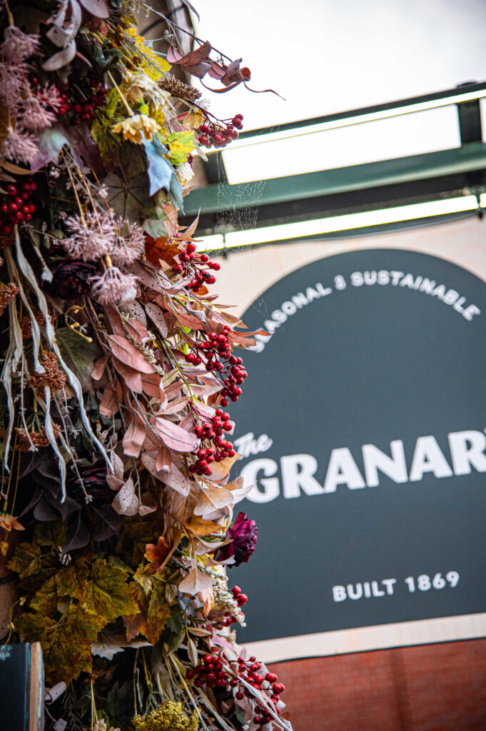 The Granary's dark green sign reading "Seasonal & Sustainable, The Granary, Built 1869", framed by dried flowers