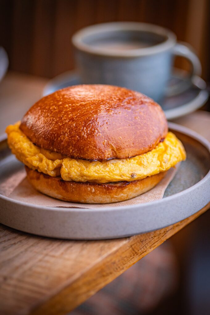 A brioche breakfast bun filled with egg on a grey plate, with a cup of coffee behind, at The Granary
